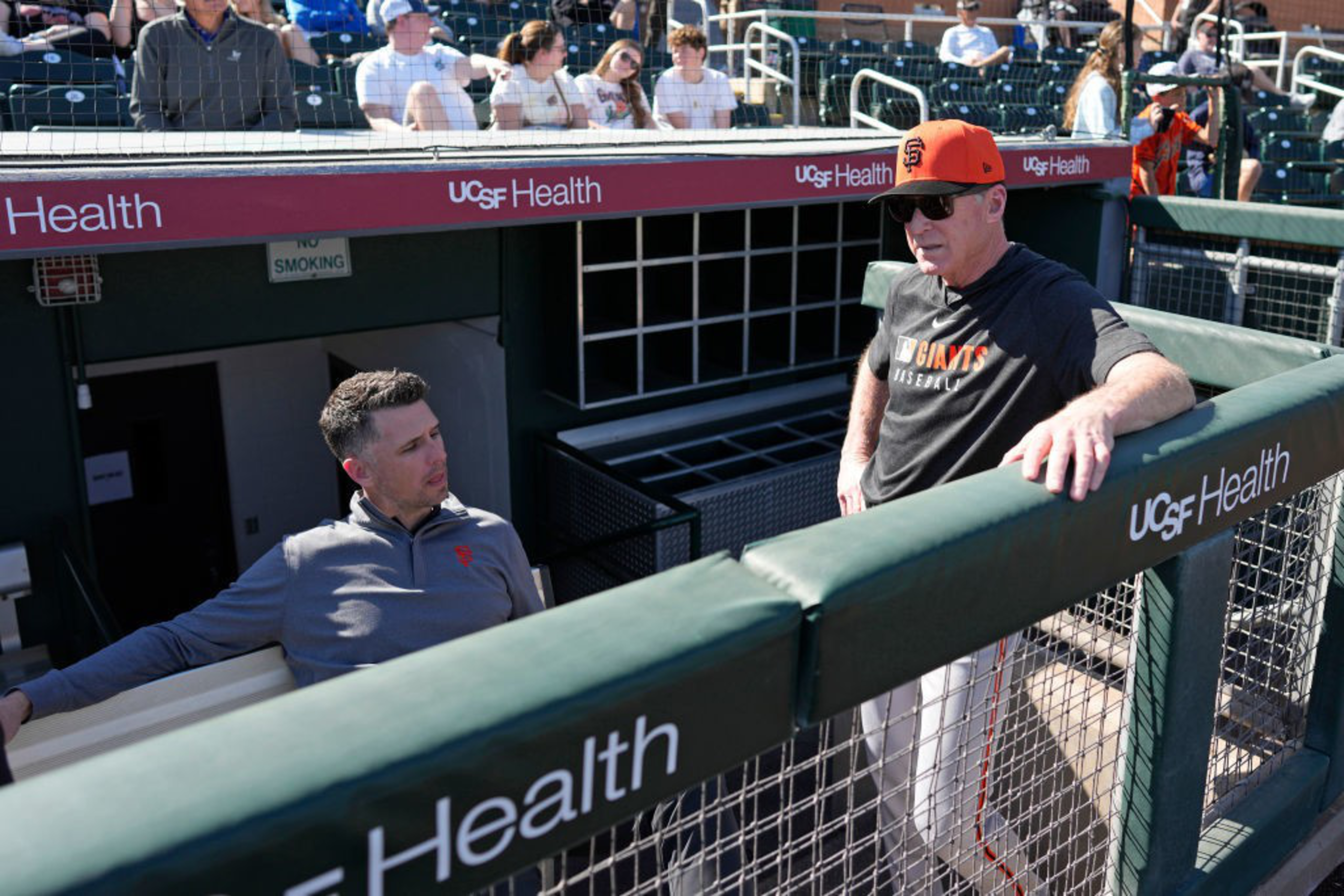 Two men are in a baseball dugout; one is seated wearing a zip-up jacket, and the other stands with sunglasses and a cap. Spectators sit in the stands behind them.