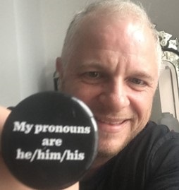 Sam Hall, local GP, holding a button that says "My pronouns are he/him/his"