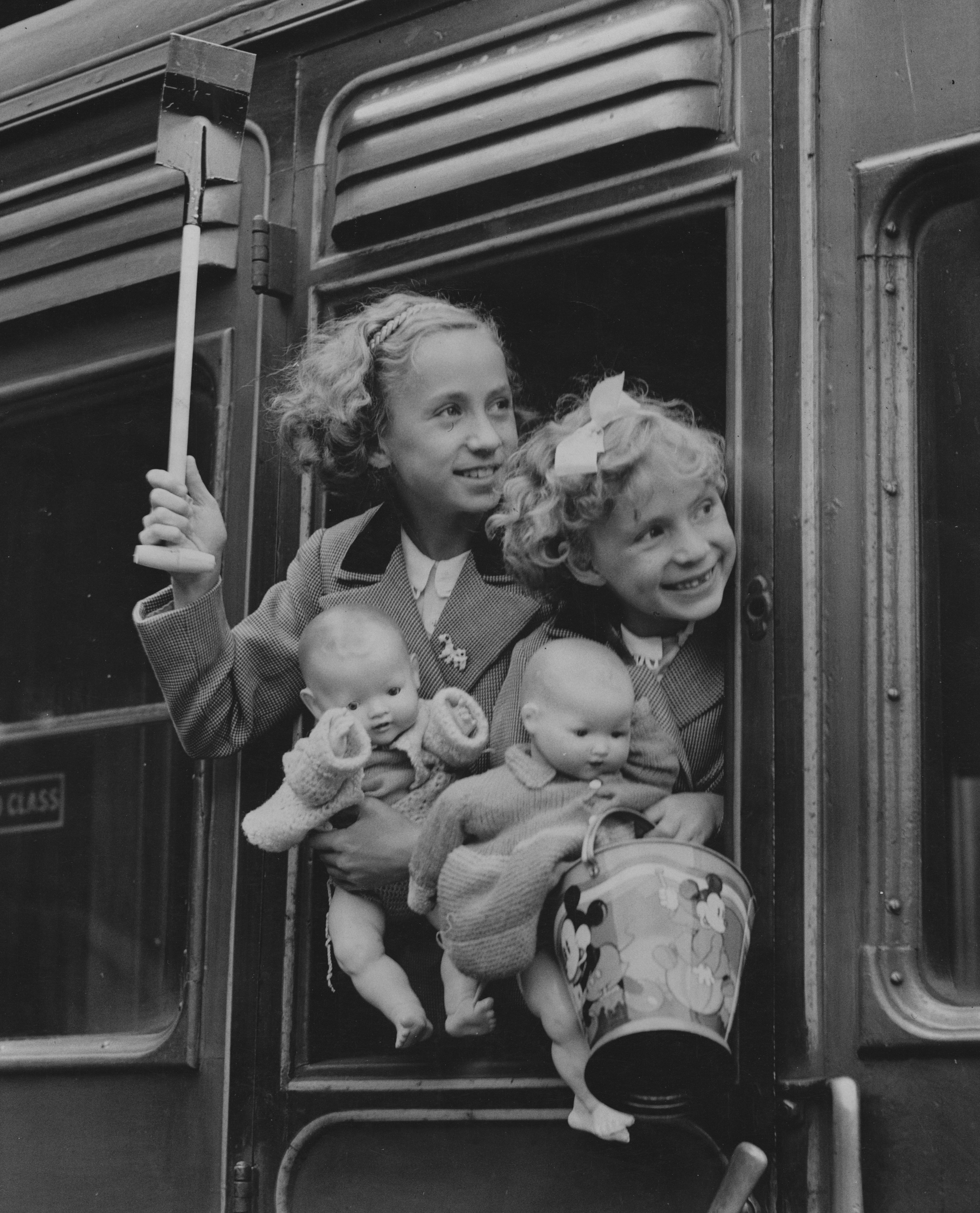 Two girls on a train, holding dolls and a bucket and spade.