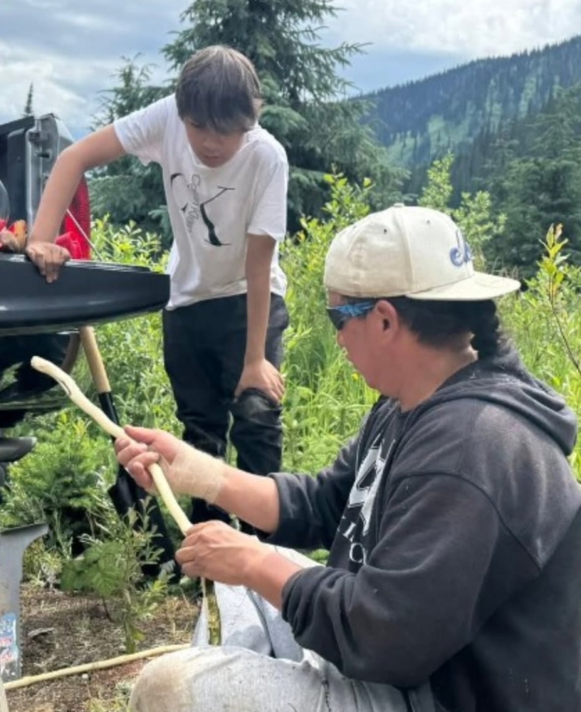 A man sits on the ground with a stick, as a younger boy watches him. A green hill can be seen in the background.