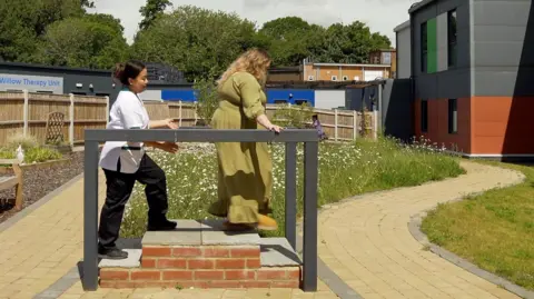 Jonny Michel/BBC Mary Hines in the garden of the West Norwich Community Hospital run by Norfolk Community Health and Care. She is with an occupational therapist and is climbing up and down a set of stairs to test how she deals with exercise. It is a new garden with brickweave pathways and a grassed area with benches