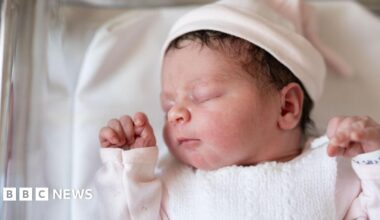 A newborn baby wearing a pink top and a pink hat sleeps in a hospital cot