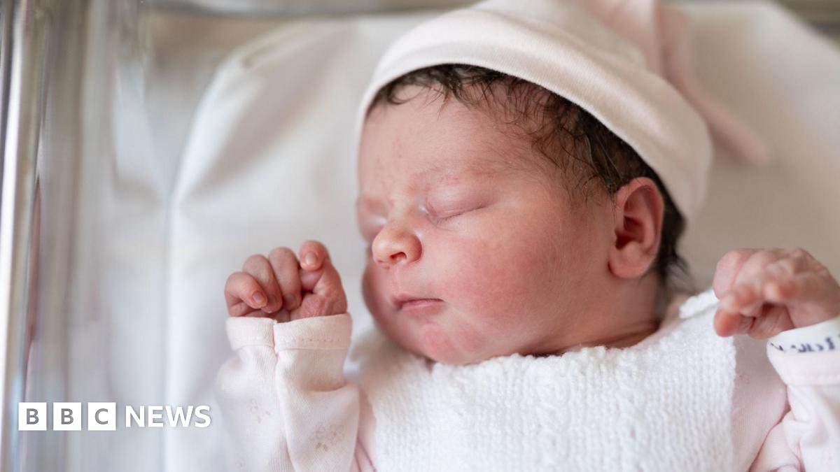 A newborn baby wearing a pink top and a pink hat sleeps in a hospital cot