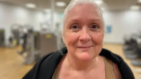 Nikki Fox/BBC Deborah Caitens-Smith standing in the main gym at Brandon Leisure Centre. She is wearing a black hoodie around her shouders and has a khaki top on underneath. She has grey hair which is tied back and is looking at the camera. She has a small piercing in her nose.
