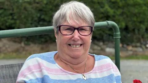 Close up image of Helen smiling looking at the camera. She is sat on a bench outside.  Helen is wearing a long sleeve top with white, blue and pink stripes. She has short grey hair and wears purple glasses. It is a head and shoulders shot of her.