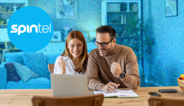 A young couple sitting at a table and cheering while looking at a laptop. The background is blue and the SpinTel logo is in the top left.