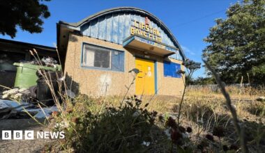 The Lifstan Boys Club building, surrounded by overgrown grass and weeds, with an overflowing green bin to the side. The building has a pebble-dashed front with two blue-framed windows and a yellow front door. The upper half of the building is a domed curved roof with blue metal fronting and the words Lifstan Boys Club in yellow writing.
