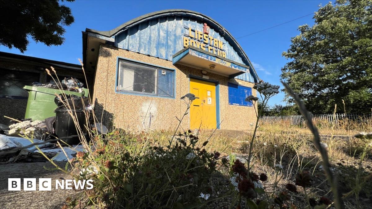 The Lifstan Boys Club building, surrounded by overgrown grass and weeds, with an overflowing green bin to the side. The building has a pebble-dashed front with two blue-framed windows and a yellow front door. The upper half of the building is a domed curved roof with blue metal fronting and the words Lifstan Boys Club in yellow writing.