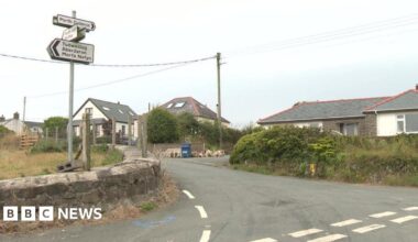 A wide image of the junction into Llangwnnadl village. On the left there are road signs attached to a pole. On the right of the image are houses.