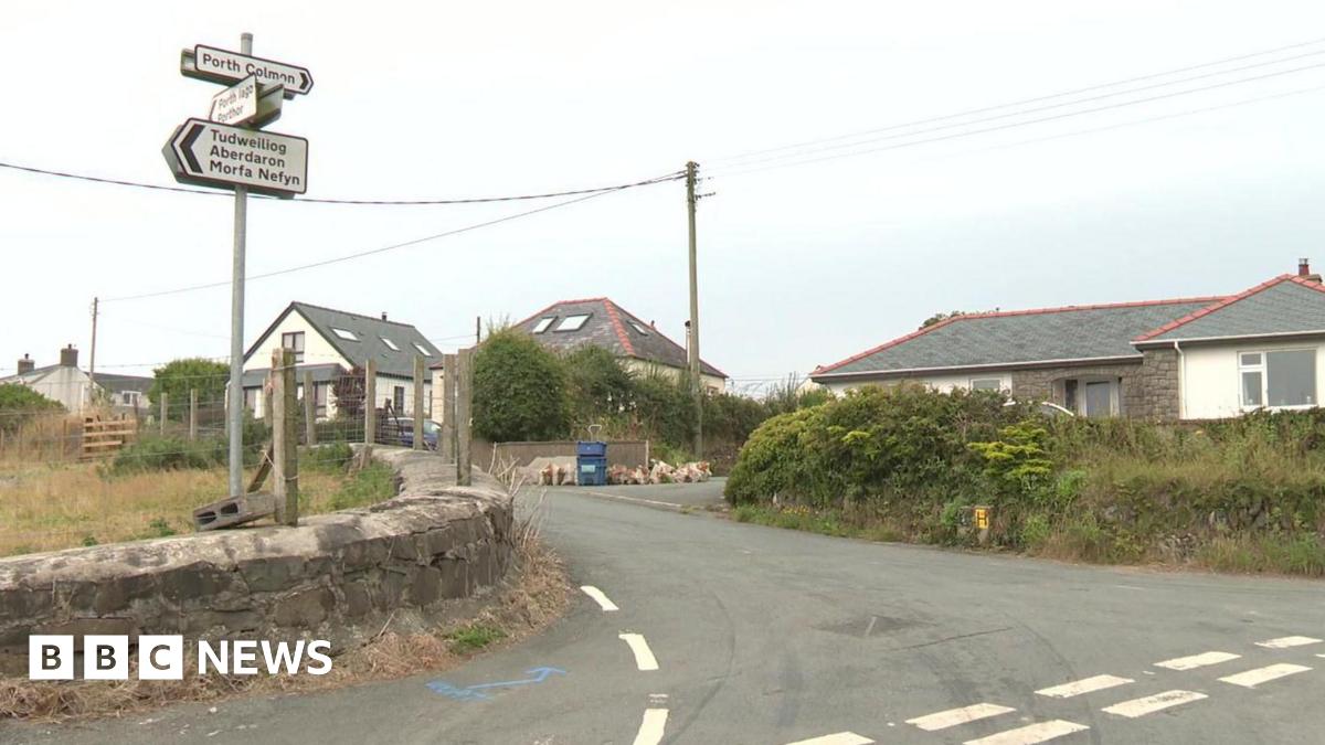 A wide image of the junction into Llangwnnadl village. On the left there are road signs attached to a pole. On the right of the image are houses.