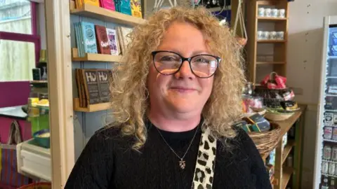 BBC Cherylee Barker smiles at the camera. She is stood in a cafe which shelves and chocolate bars in the background,