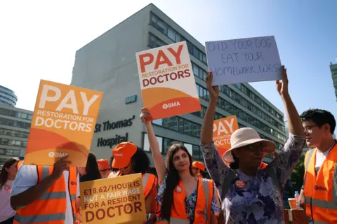 EPA/Shutterstock A group of resident doctors on strike hold orange placards calling for pay restoration outside St Thomas's hospital in London, which is visible behind them. 