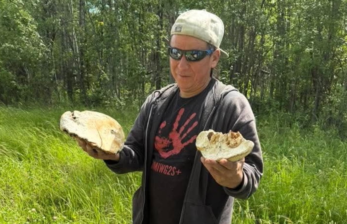 A man in a backwards ball cap and sunglasses stands with his arms outstretched, holding something in either hand.