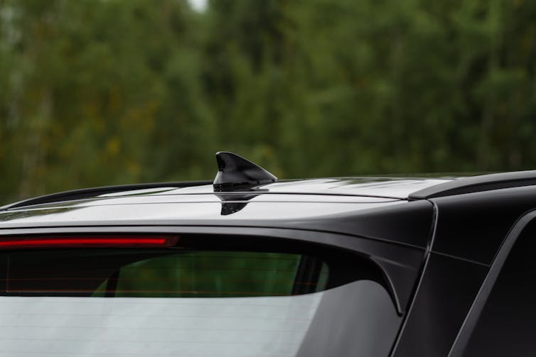 Top part of black car back showing shark finn antenna with trees in the background