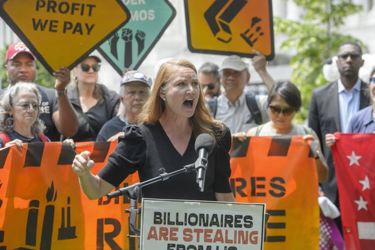 A woman speaks outdoors in front of a microphone as several peopple holding a banner stand behind her.