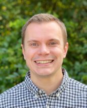 Headshot of man wearing a plaid shirt standing in front of greenery.