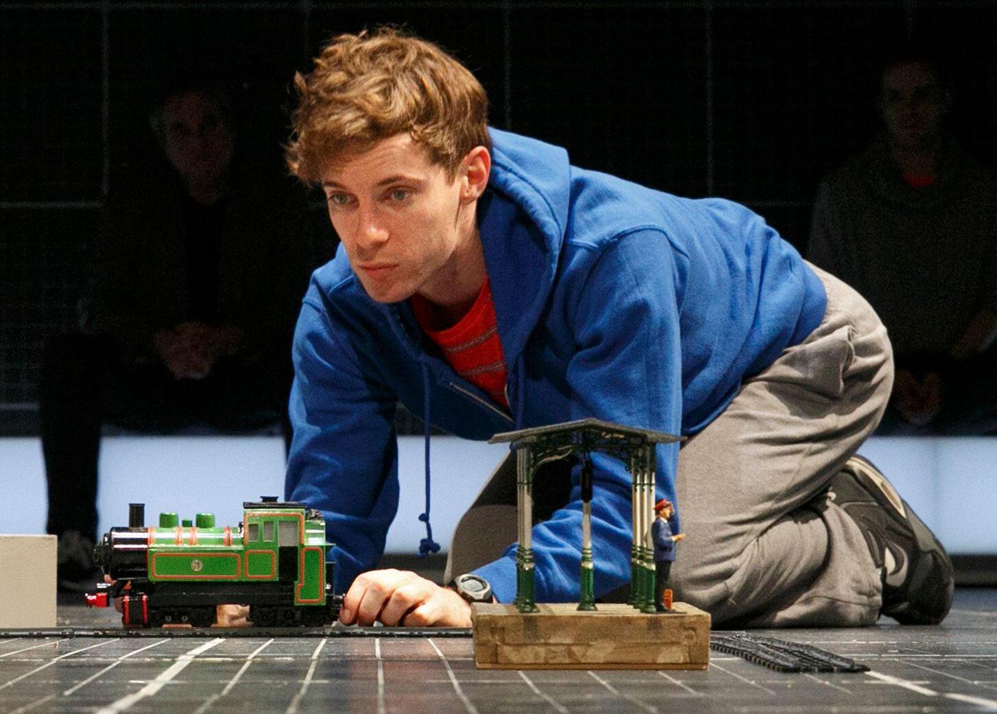 Luke Treadaway playing Christopher Boone in a stage production of *The Curious Incident of the Dog in the Night-Time*, interacting with a model train set.