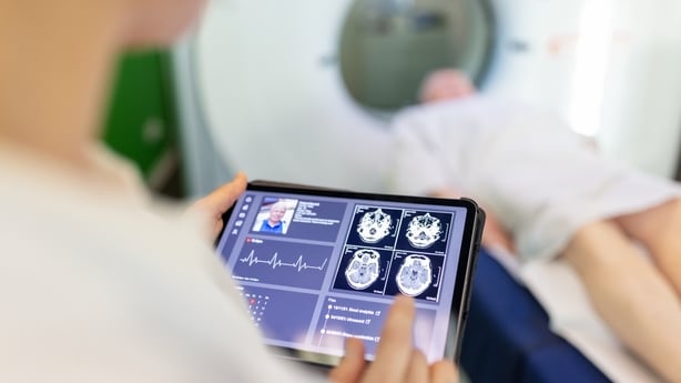 Over the shoulder view of a female doctor looking at brain x-ray images of male patient undergoing MRI in background at hospital. Neurologist examining patient's CAT scan images on digital table in clinic.