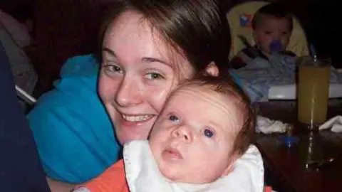 Becci Smart Becci Smart holding Zak when he was a baby - she is behind him in a blue top and has her dark hair pulled back in a ponytail and has a big smile on her face. Zak is a young baby with an orange top on and a white bib over the top, he has very short fluffy baby hair and is looking up at the roof. There is a baby in a high chair with a dummy in his mouth seen in the background of the image.