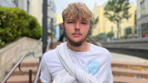 BBC Boy with a short hair but a long blonde fringe and beard looking at the camera. He is wearing a white t-shirt with a blue logo on the right and a white jumper tied over his shoulder.