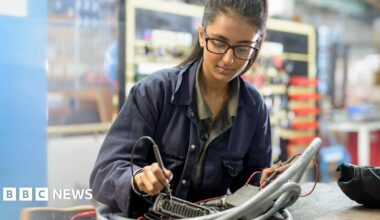Stock photo of an employee with long hair and glasses working on a soldering desk in an engineering environment while wearing work overalls.