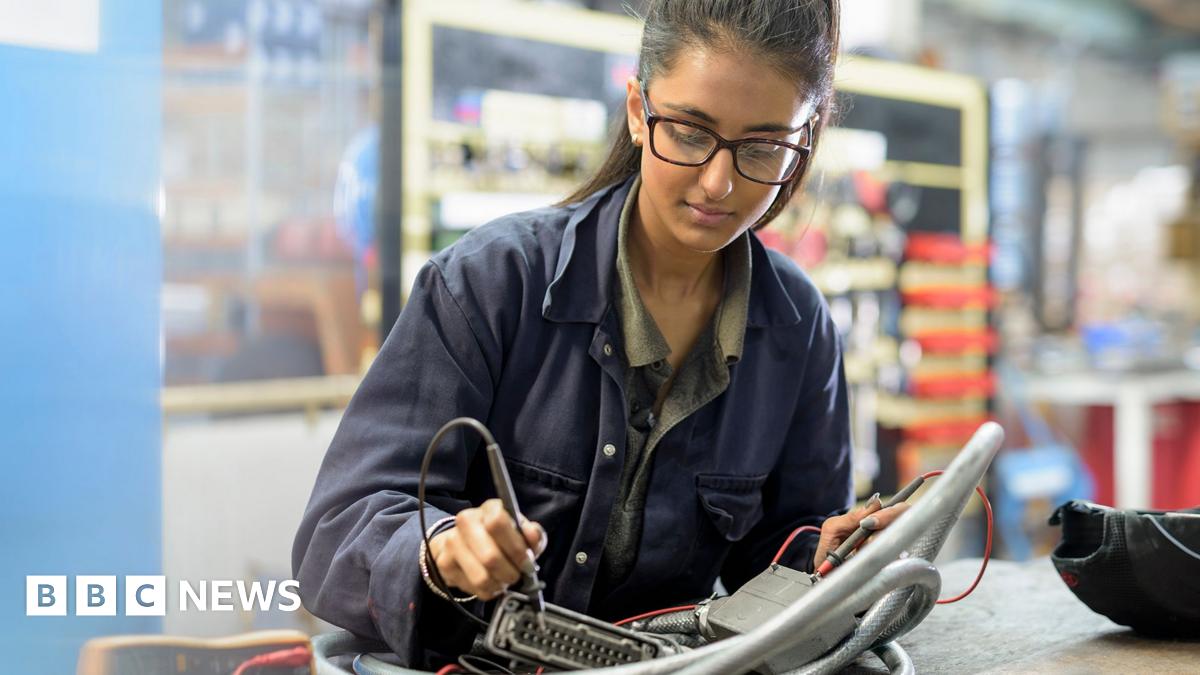 Stock photo of an employee with long hair and glasses working on a soldering desk in an engineering environment while wearing work overalls.