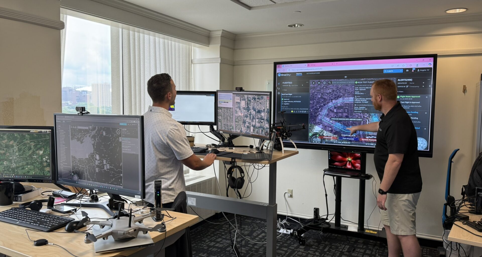 Two men stand in an office looking at four smaller computer screens and one larger computer screen. There is a tan desk in the foreground housing two computer monitors and a small grey drone. In the background stands a man in a white polo shirt facing two other computer monitors on a desk. Behind this set up is a large TV monitor with a man standing with his arm reached out in front. The man is wearing a black polo shorts and tan shorts as well as white tennis shoes. The screen shows a map and different pieces of software working in unison.