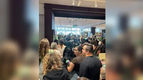 Amy White A picture inside a large bookshop at the back of a large crowd, which stretches to the far, windowed wall.