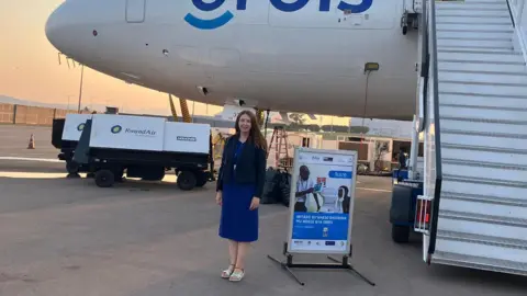 Orbis A dark-haired woman in a blue dress and jacket stands in front of a passenger jet plane.