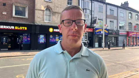 BBC A man wearing a light blue T-shirt and black-framed glasses looking at the camera. He is standing in a city street. On the other side of the road is a Merkur Slots arcade and a number of fast-food restaurants.
