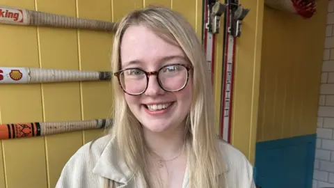 Grace Pickett A woman with long blonde hair and brown glasses in sat inside a building with yellow walls. She is smiling at the camera