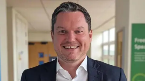 Gloucestershire Hospitals NHS Foundation Trust A picture of Kevin McNamara in a corridor, he is wearing a navy blazer and a white shirt, and smiling widely.