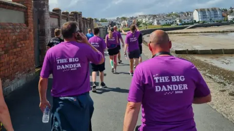 Brian Elwood A group of people in purple T-shirts with white lettering on the backs reading "The Big Dander". They are walking along a seaside promenade, a beach is to the right and a red brick wall to the left. The sky is blue with a few clouds.