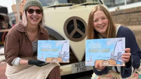 Sunderland BID Jo Gordon and Hannah Graham holding a copy of The Story of the Lazy Loaf and smiling as they kneel in front of a cream-coloured van. The book cover is blue and shows bread loaves surrounded by Sunderland landmarks, such as Roker Pier and Fulwell Windmill. Jo has light pink-coloured hair, wears a green beanie, sunglasses, brown trousers and jacket. Hannah has long straight strawberry blonde hair and is wearing a blue cardigan.