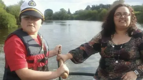 Jo Cozens Tomas Cozens as a young boy with his mum Jo. They are on a boat on a river and are holding a wooden handle that steers the boat. Tomas is wearing a red t-shirt and a black and red life jacket with a white, Navy-style captain's hat. His mum has curly dark hair and rectangle-shaped glasses with a dark, patterned long-sleeved top on. The river and trees are visible in the background 