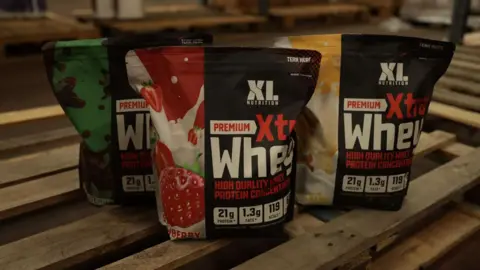Jordan Dales Three bags full of XL Nutrition Whey protein on a wooden pallet. On the black bags which are sealed at the top there is a photo of a strawberry, another has a banana and another is chocolate mint. It says Premium Xtra Whey on each bag.
