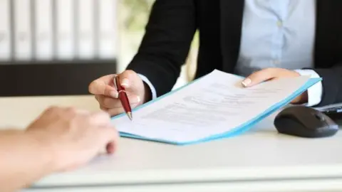 Getty Images a woman wearing a blue shirt and black blazer hands a piece of paper and pen across a desk to another person sat opposite her. A computer mouse and keyboard are visible at the edge of the desk 