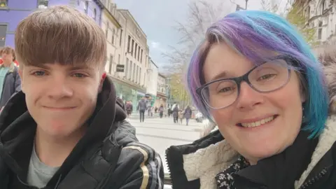Becci Smart Becci Smart taking a selfie with Zak on a pedestrianised city centre street. Zak is on the left and his light brown hair is brushed forward so his fringe covers his forehead. He has a light grey t-shirt on underneath a black Adidas puffa jacket. Becci has a bob-cut hairstyle which is purple and blue. She is wearing blue-rimmed glasses and a black coat over a black dress with white flowers on. People can be seen walking down the street behind them between various shop fronts. It is a head and shoulders shot of the two of them.