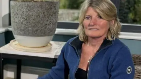 A woman sits wearing a blue top in a room with two windows and a large vase behind.
