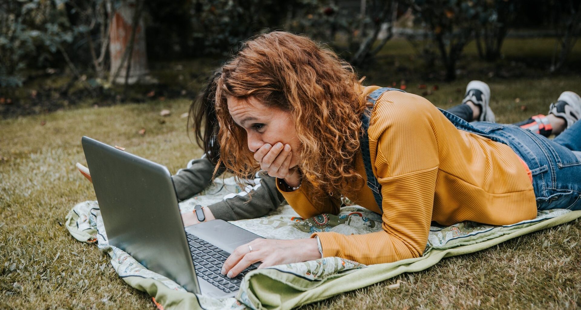 A mother and daughter using a laptop while laying on a blanket in their backyard.