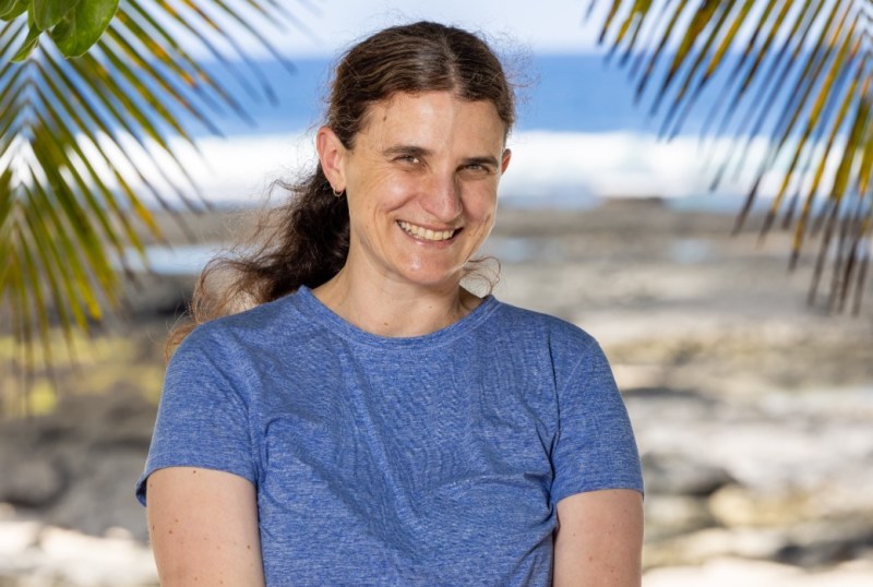 Smiling woman in a blue t-shirt stands outdoors with a beach in the background, surrounded by palm leaves, showcasing a sunny day in Australia.