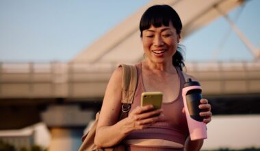 Woman holding bottles of electrolytes, smiling and looking at phone while outside in hot weather