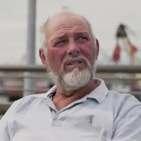 Rob Caunter, 66 years old, is seated and staring off camera to the right of frame. He is sitting down on a bench in the port with fishing boats behind him. He has white hair and a white beard and is wearing a grey and white polo T-shirt.