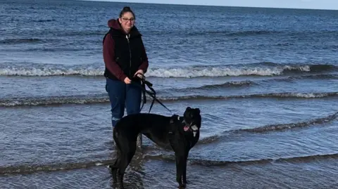 Handout A photo of Lotte stood with her dog Tina, a black whippet, in the shallow waves of a beach. She has walking boots, blue jeans rolled up, and is wearing a dark gilet over a maroon hoodie.