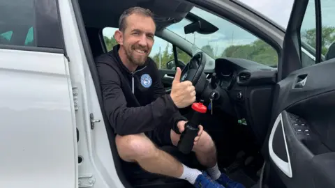 PA Media Camera Alexander sitting in the driving seat of a white vehicle with the door open. He is in sports gear and is holding a black water bottle and smiling while holding his thumb up.