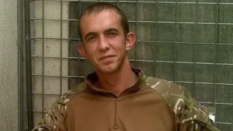 PA Media A grainy headshot of Camera Alexander wearing a camouflage Army jacket. He is standing in front of a metal fence and looking into the camera.