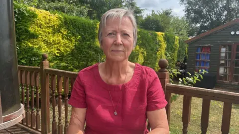 BBC A woman in a dark pink top and grey hair sitting at a garden table
