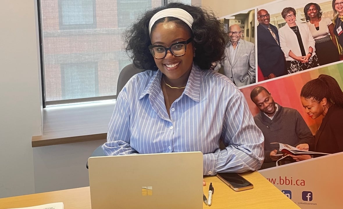 Woman wears blue collared shirt and sits at desk with laptop. 