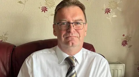 A man sitting on a red leather sofa wearing glasses, a white shirt and a brown and grey striped tie