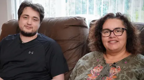 Tomas Cozens and his mum Jo sitting next to each other on a brown leather sofa. Tomas is on the left and is wearing a black Under Armour t-shirt and has dark wavy hair and a brown goatee beard. Jo has curly brown hair down to her shoulder and has a green floral print top on that has laces on the chest. It is a head and shoulders shot of them.